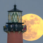 The full moon rises behind the Jupiter Inlet Lighthouse. Guests stand at the top of the lighthouse and look into the distance.
