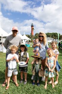 A mother, father, and their five children stand in a green field and smile at the camera. The Jupiter Inlet Lighthouse stands behind them.