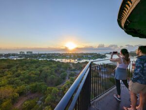 Two guests stand at the top of the Jupiter Inlet Lighthouse. They face east as they watch the sun rise. One takes a picture of the sunrise.
